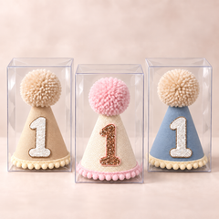 Three first birthday party hats in clear boxes in tan, pink and blue, each with a glitter number one